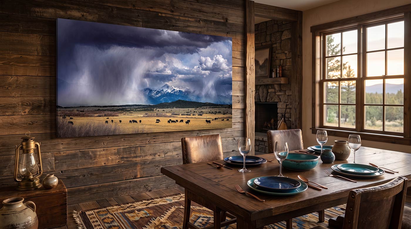 A large wall-mounted canvas print of a dramatic mountain landscape with a stormy sky and snow-capped peaks, displayed in a rustic dining room with a wooden table, chairs, and window.