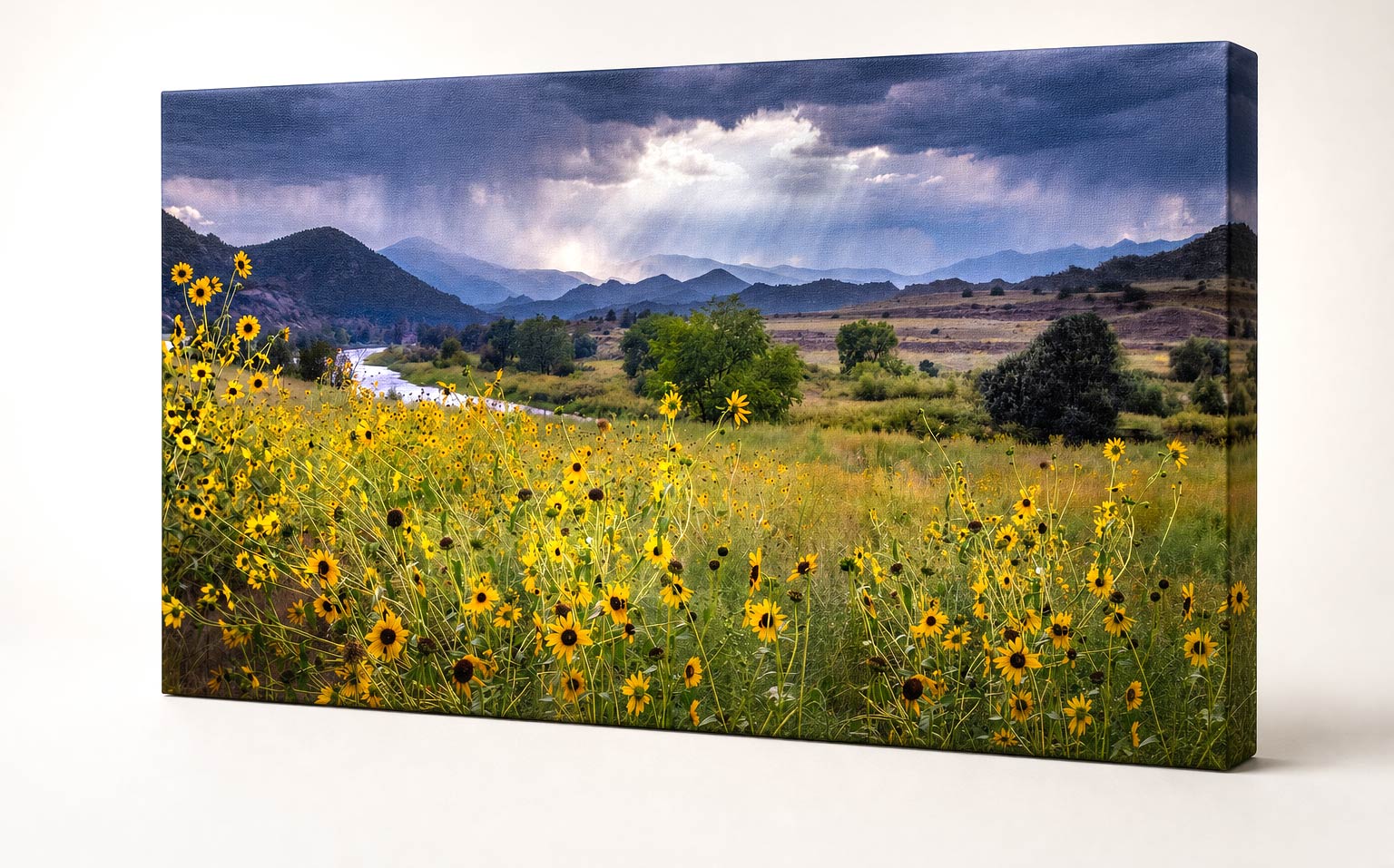 A rectangular canvas wall art print showing a scenic landscape with a yellow wildflower meadow in the foreground, a winding river, distant blue mountains, and dramatic storm clouds with light rays breaking through. The canvas is photographed at a slight angle against a clean white background.