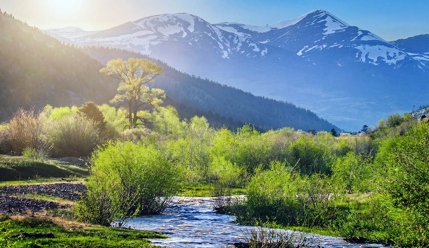 A scenic landscape print featuring a winding stream in a lush green valley, with snow-capped mountains and a bright sun in the background.
