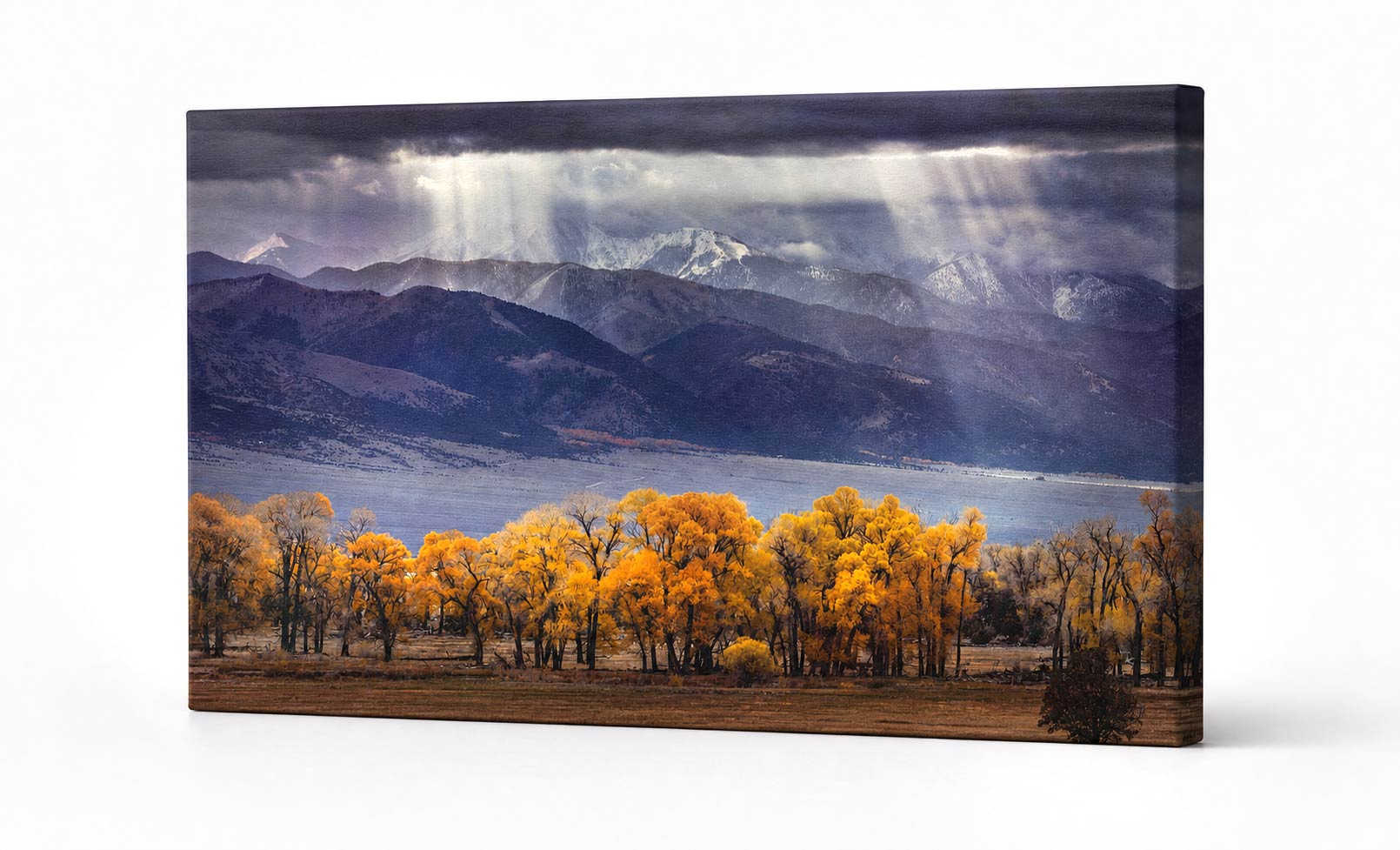 A rectangular canvas wall art print showing a dramatic mountain landscape with golden yellow trees in the foreground, a lake, and sunbeams breaking through dark clouds. The canvas is photographed at a slight angle against a clean white background.
