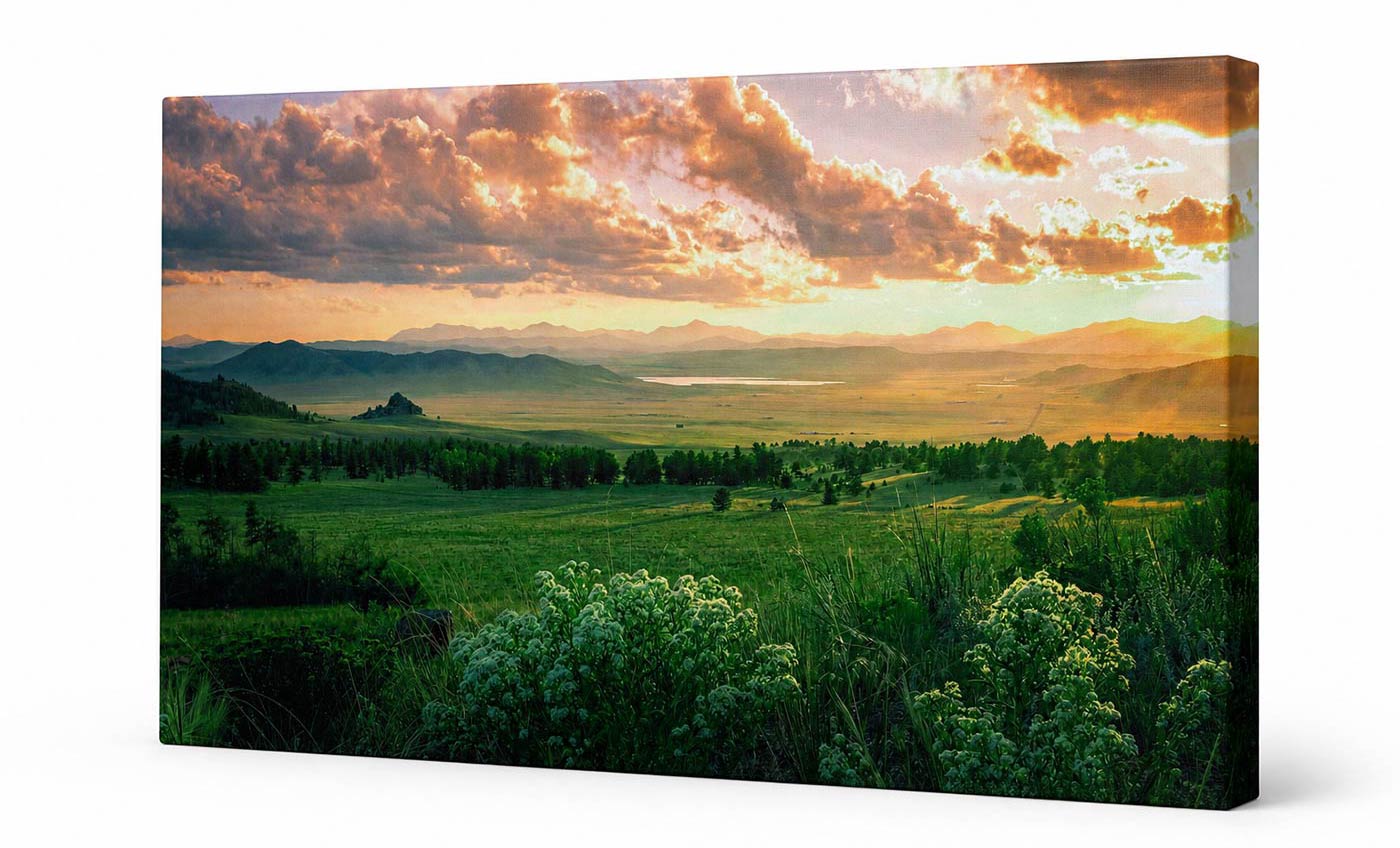A rectangular canvas wall art print showing a scenic landscape with green fields, distant mountains, and a dramatic sunset sky filled with orange, gold, and dark clouds. The canvas is photographed at a slight angle against a clean white background.