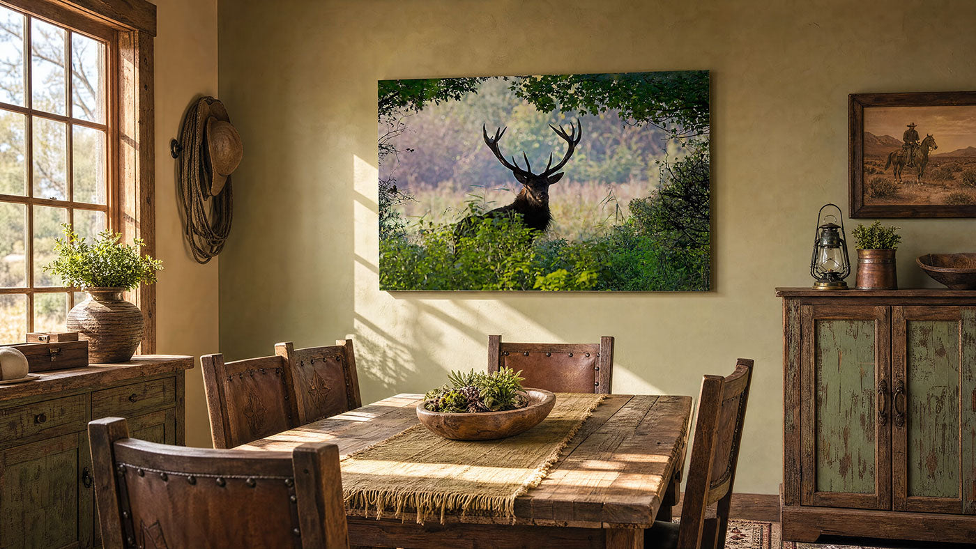 A large rectangular wall art print showing a deer in a forest hangs on a beige wall in a rustic dining room with a wooden table, chairs, cabinet, and window.