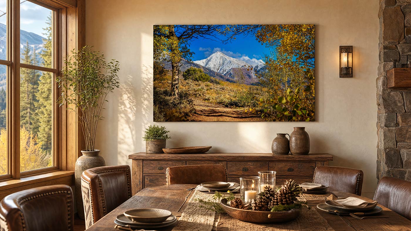 A large rectangular wall art print depicting a mountain landscape with autumn trees and a blue sky, displayed above a wooden dining table in a rustic dining room.
