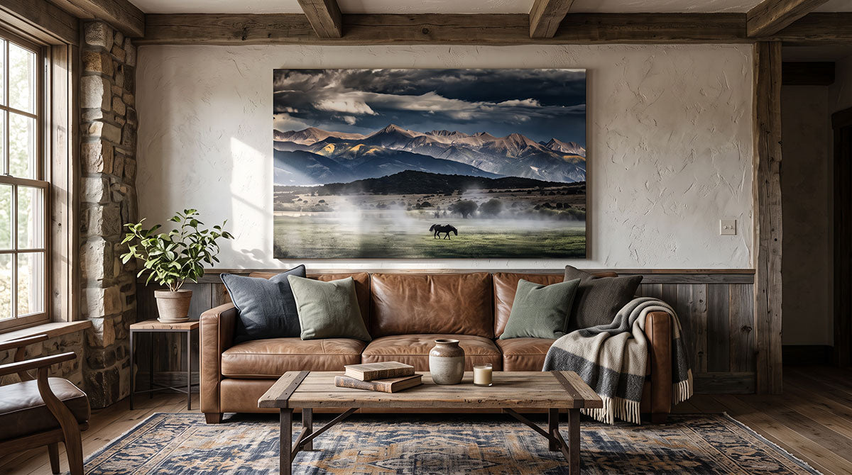 A large rectangular canvas print of a mountain landscape with mist, clouds, and a small horse, displayed on a wall above a brown leather sofa in a rustic living room.