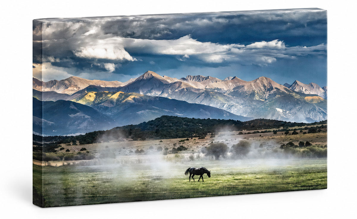 A rectangular canvas wall art print showing a dramatic mountain landscape with a horse in a misty green valley under a cloudy blue sky. The canvas is photographed at a slight angle against a plain white background.