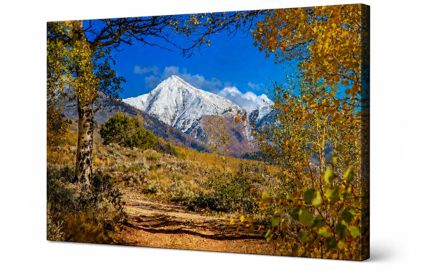 A rectangular canvas wall art print showing a mountain landscape with a snow-capped peak, blue sky, autumn trees, and a dirt path. The canvas is photographed at a slight angle against a plain white background.