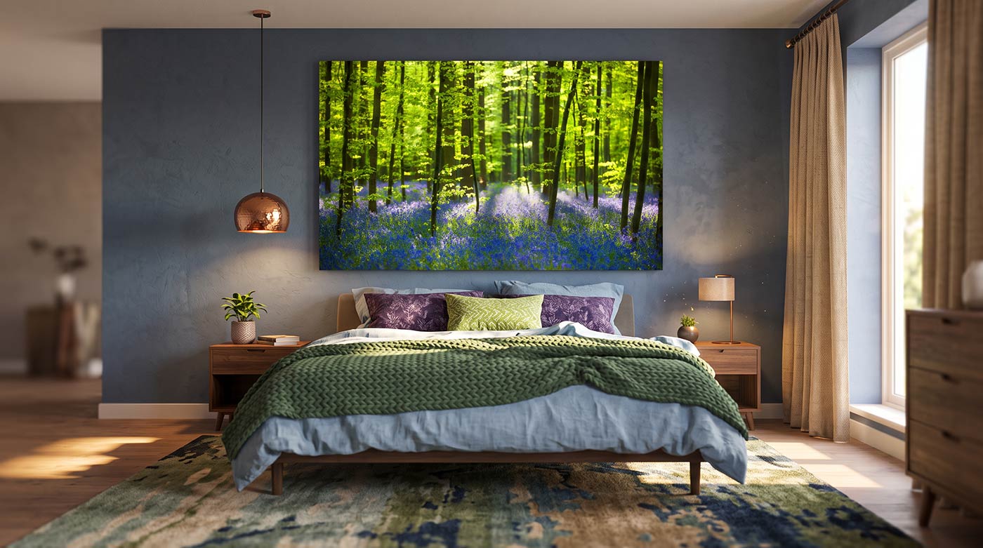 A large square canvas wall art print depicting a vibrant green forest with bluebells, hung on a textured slate blue-gray wall above a bed in a modern styled bedroom.